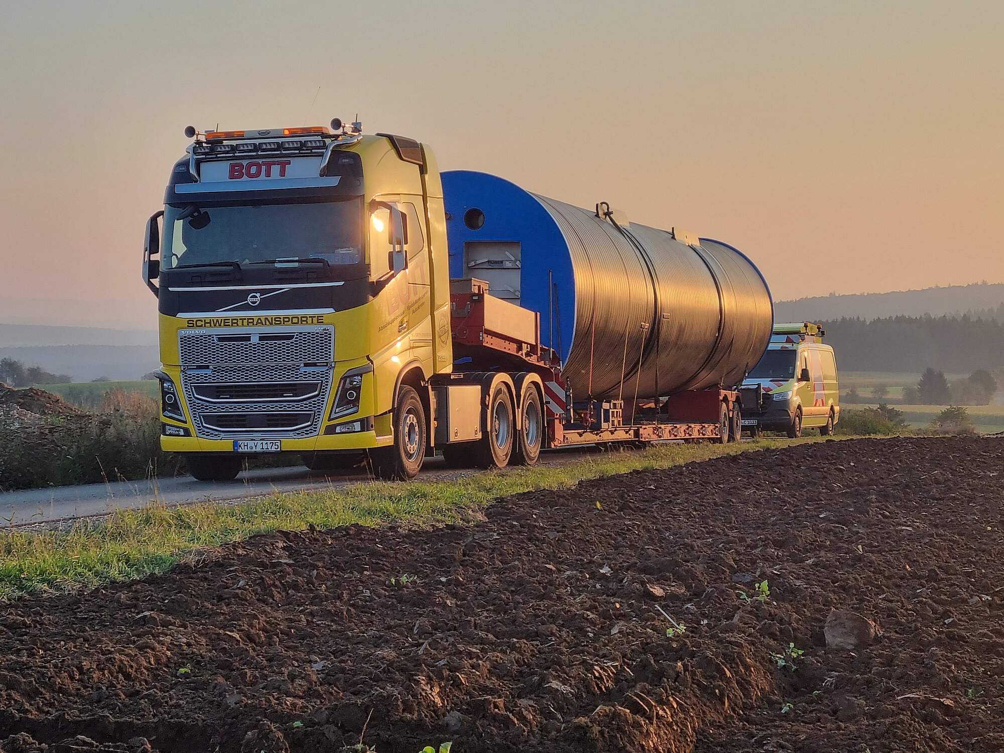 Gelber Lastwagen mit großer Ladung und Begleitfahrzeug auf ländlicher Straße unterwegs